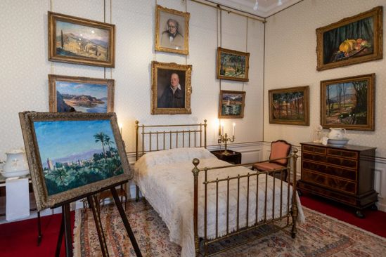 Bed in centre of room next to painting on easel, white walls with many gold-framed paintings