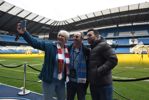 Three people taking selfie in front of Manchester City Stadium pitch