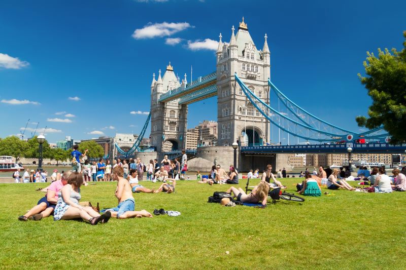 People sat enjoying sunny day on green grass besides Tower Bridge in London