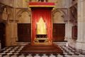 Coronation Chair at Westminster Abbey in centre of room with balck and white checkered floor, chair is painted slightly gold and is glowing from spotlight